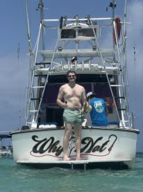 A man stands on the back of the 'Why Nat' boat, enjoying a sunny day on the water, showcasing a fun and relaxed atmosphere perfect for group outings.