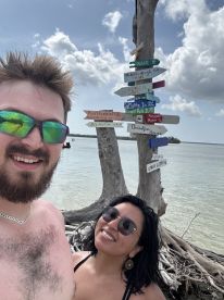 A cheerful couple poses for a selfie at a scenic beach with directional signs in the background, capturing the essence of their fun vacation adventure.