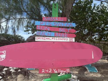 A colorful sign at a beach location, highlighting activities like pig swimming and sunset views — a fun destination for visitors.