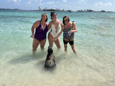 Three women enjoy a playful encounter with a swimming pig in shallow waters, creating a memorable wildlife experience in the Bahamas.