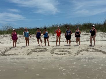 A lively group of women celebrates on the beach, posing with drinks beside the sand art spelling 'LPGA' — perfect for memorable beach outings and celebrations.