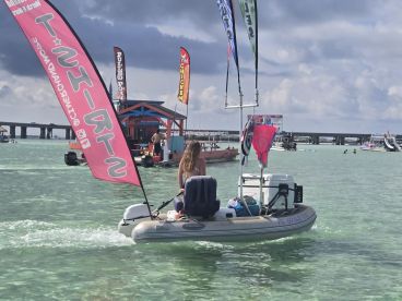 A woman navigates a small boat in clear waters, showcasing a vibrant atmosphere with nearby vendors and activities — perfect for a fun day at the beach.