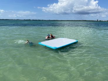 A group enjoys a fun day in the water, with one person snorkeling and others relaxing on a floating mat — a perfect way to experience the beauty of the ocean.