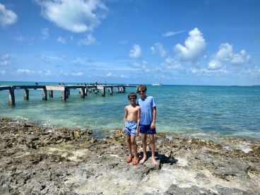 Two boys pose on the rocky shore with a picturesque pier and sailboat in the background, capturing a fun family moment by the ocean.