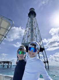 Two boys pose happily on a boat with a lighthouse in the background, capturing a joyful moment during their ocean adventure.