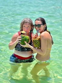 Two friends enjoy refreshing cocktails served in watermelons and pineapples while wading in crystal-clear waters, capturing the essence of a fun day on the beach.