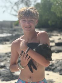 A young boy smiles while holding a sleeping pig on a beach, showcasing a unique and playful interaction with wildlife in a tropical setting.