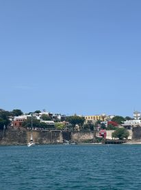 A picturesque view of a coastal harbor with a sailboat anchored near a historic fortification, showcasing the vibrant architecture and lush greenery of the area.