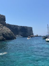 A picturesque view of boats navigating through crystal-clear waters near dramatic cliffs, with a swimmer enjoying the refreshing sea — a perfect snapshot of a vibrant day on the water.