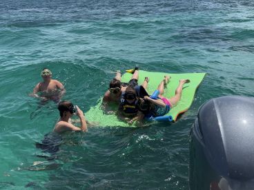 A lively group enjoys a fun day in the water, engaging in snorkeling and relaxing on a floating mat — perfect for summer adventures and family outings.