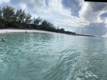 A group enjoys a refreshing swim in the crystal-clear waters near a sandy beach, with a boat anchored in the background — perfect for a fun day at the sandbar.