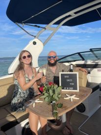 A couple celebrates their engagement aboard a boat, raising glasses in a toast with a beautiful bouquet and a sign marking the special occasion.