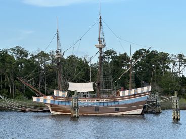 A beautifully crafted ship docked along the shore, showcasing its intricate rigging and classic design against a serene backdrop of trees and sky.