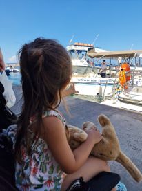 A young girl waits at the marina, holding her stuffed animal while watching the boat 'Serenity Boat Charters' — a charming moment capturing the excitement of a day on the water.