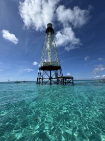 A stunning view of a lighthouse standing tall over crystal-clear waters, with boats nearby and people enjoying the beautiful scenery — a perfect destination for water activities.