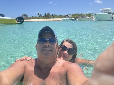 A couple enjoys a refreshing swim in crystal-clear waters, capturing a joyful selfie with boats anchored in the background — a perfect moment for beach lovers.