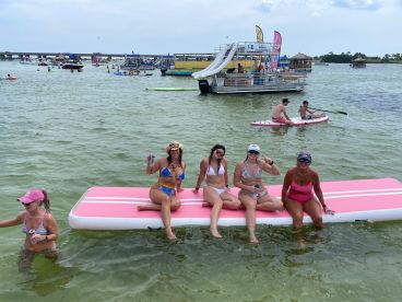 A lively group enjoys a fun day on the water, relaxing on a floating mat while others paddleboard nearby — perfect for summer parties and water activities.