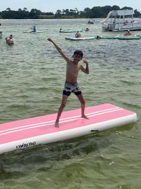 A young boy joyfully poses on a pink floating mat while enjoying a day of water activities, surrounded by friends and family in a lively aquatic setting.