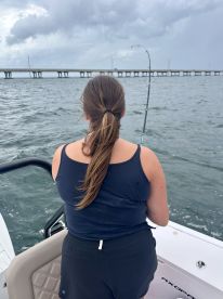 A woman enjoys fishing from a boat, casting her line into the ocean while a bridge looms in the background — a perfect snapshot of a day on the water.