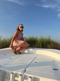 A woman poses on the deck of a boat against a backdrop of tall grass and a clear sky, capturing a serene moment on the water.
