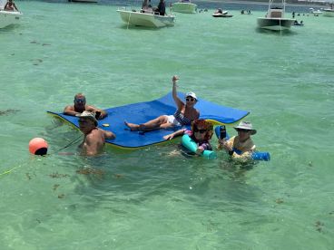 A lively group enjoys a fun day in the water on a floating mat, celebrating with drinks and laughter in a beautiful aquatic setting.