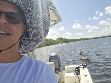 A man smiles for a selfie on a boat while a pelican perches nearby, showcasing a delightful moment of wildlife interaction on the water.