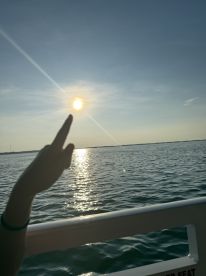 A hand points towards the sun reflecting on the water, capturing a serene moment during a sunset cruise — perfect for those seeking relaxation on the water.