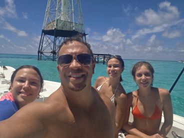 A joyful group of friends poses for a selfie on a boat near a historic lighthouse, enjoying a sunny day on the water — perfect for memorable boating adventures.