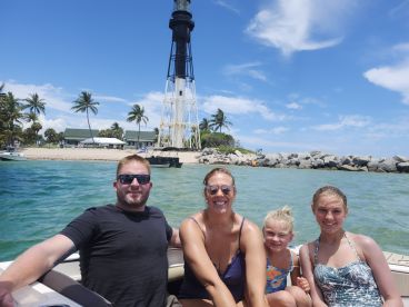 A joyful family poses on their boat with a scenic lighthouse in the background, capturing a perfect day on the water.