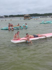 A lively group enjoys a fun day on the water, lounging on a paddleboard and swimming in the warm shallows — perfect for summer adventures.