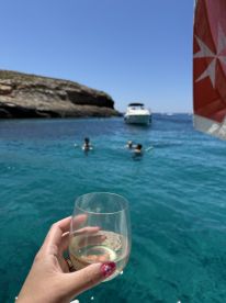 A hand holding a glass of wine captures a vibrant scene of friends snorkeling in crystal-clear waters, with a boat anchored nearby — perfect for a fun day out on the water.