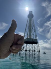 A hand giving a thumbs-up gesture in the foreground with a distant lighthouse in the background, but the image lacks clarity.