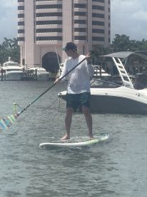 A man paddleboards in calm waters, showcasing an active water sport experience against a backdrop of luxury boats and a high-rise building.