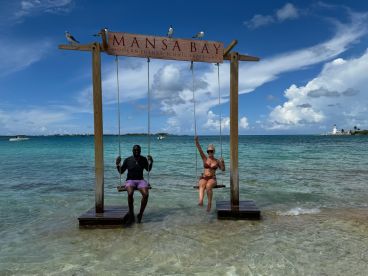 A couple enjoys a playful moment on swings at Mansa Bay, surrounded by stunning turquoise waters — a perfect spot for relaxation and fun in the sun.