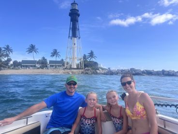 A joyful family poses for a photo on their boat with a picturesque lighthouse in the background, capturing a memorable day on the water.