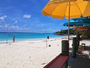 A picturesque beach scene featuring a couple walking along the shore, with a volleyball net set up against a backdrop of turquoise waters and vibrant umbrellas — perfect for a tropical getaway.