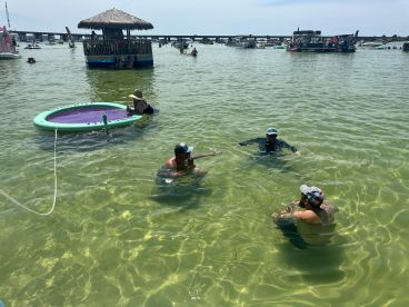 A lively group enjoys a sunny day swimming and relaxing on a floating mat in shallow waters, perfect for summer fun and social gatherings.