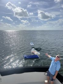 A man gestures towards a line of colorful floating mats in the shimmering water, showcasing a fun day on the boat — perfect for relaxing and enjoying the sun.
