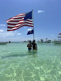 A couple stands in shallow, clear waters, proudly displaying an American flag while enjoying a sunny day at the beach, surrounded by boats — a perfect moment for summer celebrations.