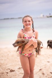 A young girl joyfully holds a piglet on the beach, showcasing a delightful interaction with wildlife — a memorable experience in the Bahamas.