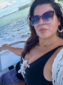 A woman enjoys a sunny day on the water, capturing a selfie on a boat with beautiful scenery in the background.