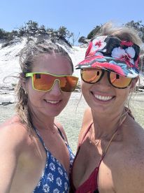 Two friends pose for a cheerful selfie at the beach, showcasing their vibrant summer styles and enjoying a sunny day by the water.