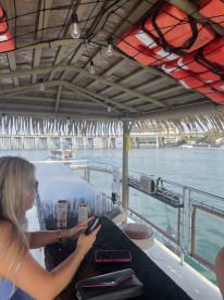 A cozy interior view of a boat's seating area, featuring a woman using her phone while enjoying drinks. The setting is perfect for relaxation and socializing on the water.