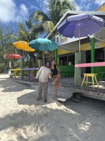 A cheerful couple poses in front of a vibrant beach bar, surrounded by colorful umbrellas and palm trees, capturing the essence of a fun day at the beach.