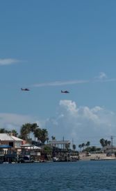 An unclear image featuring helicopters in the sky above a waterfront area, with buildings and palm trees visible in the foreground.