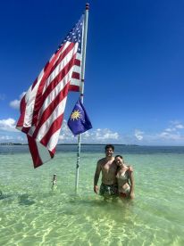 A couple enjoys a sunny day at the beach, standing in shallow water with flags waving in the background — perfect for romantic getaways or beach celebrations.