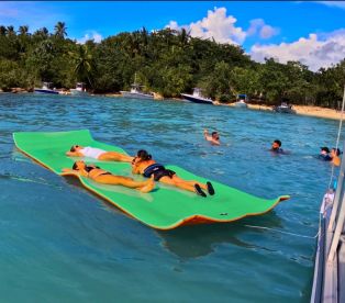 A lively group enjoys a day on the water, relaxing on a floating mat while others swim nearby — a perfect scene for summer fun and beach outings.
