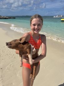 A young girl joyfully holds a pig on the beach, capturing a memorable moment of human-animal interaction in a tropical paradise.