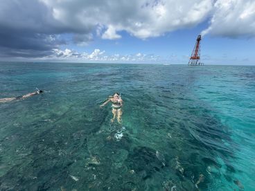 Two snorkelers enjoy the vibrant underwater scenery in clear turquoise waters, with a historic lighthouse in the background — a perfect snapshot of adventure on the water.