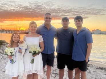 A joyful family poses for a sunset photo on the beach, celebrating a special occasion with beautiful floral arrangements and a beloved pet, creating lasting memories by the water.